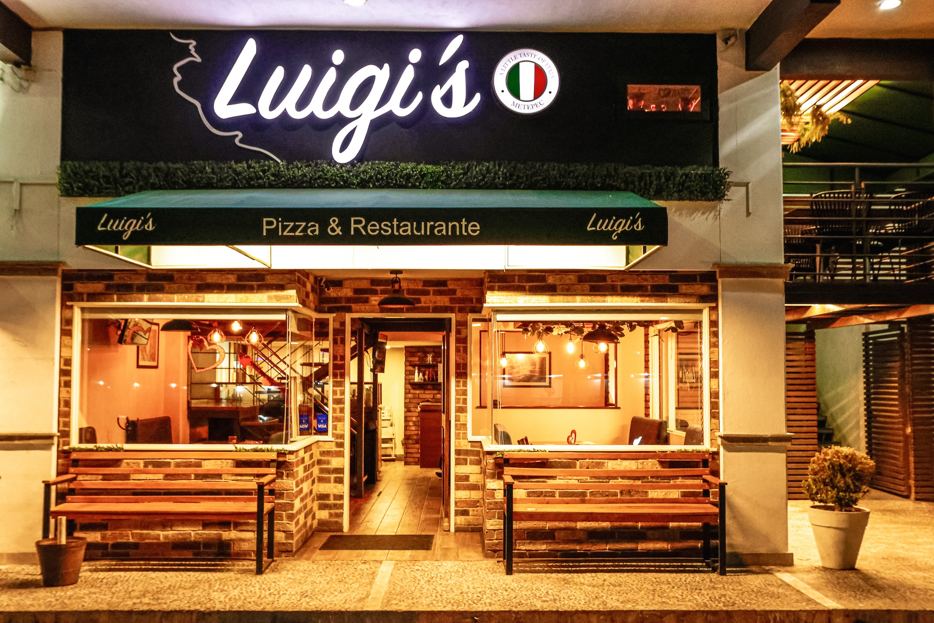 Fachada nocturna de Luigi's Pizza & Restaurante sucursal Las Torres: letrero iluminado, toldo verde y bancas de madera en la entrada